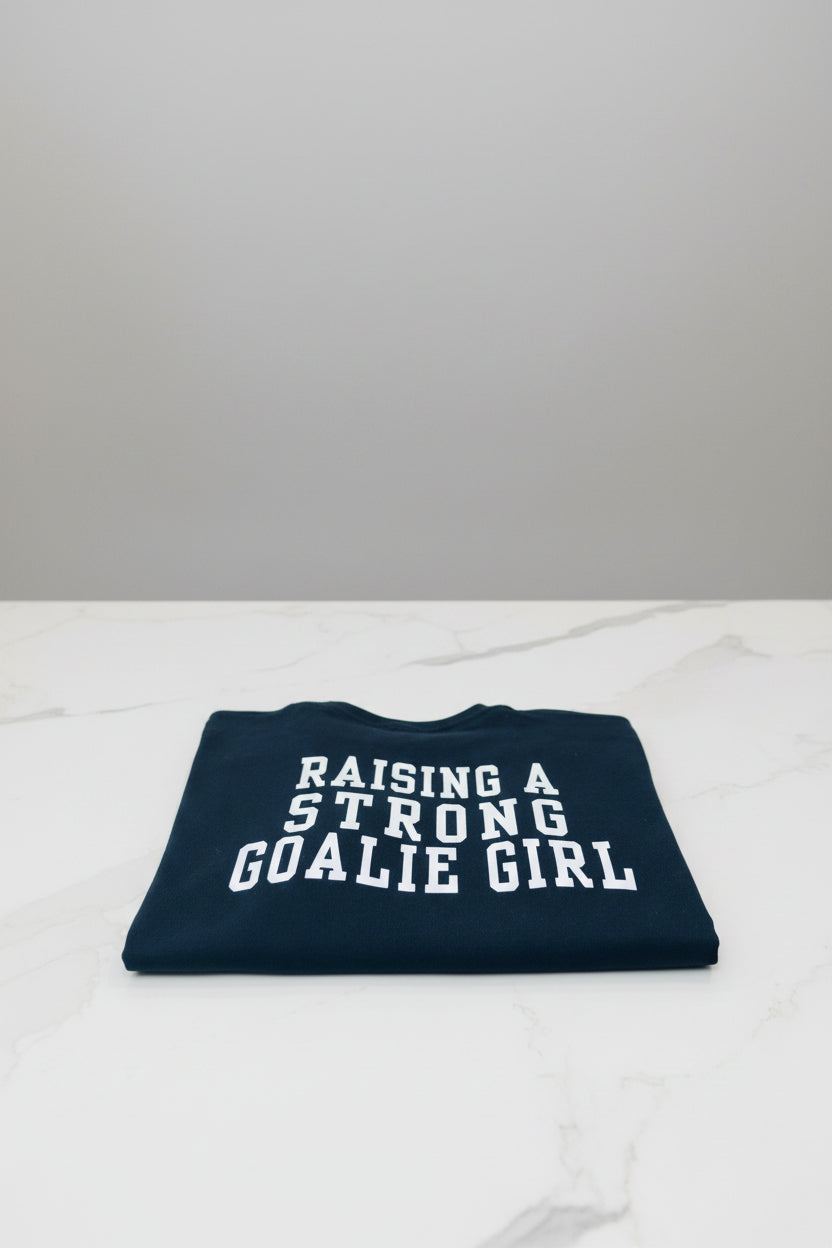 Folded black t-shirt with 'RAISING A STRONG GOALIE GIRL' text on a light background