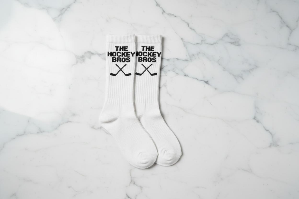 A pair of white crew socks with black 'THE HOCKEY BROS' text and crossed hockey sticks printed on the cuffs, laid flat on a white marble surface.