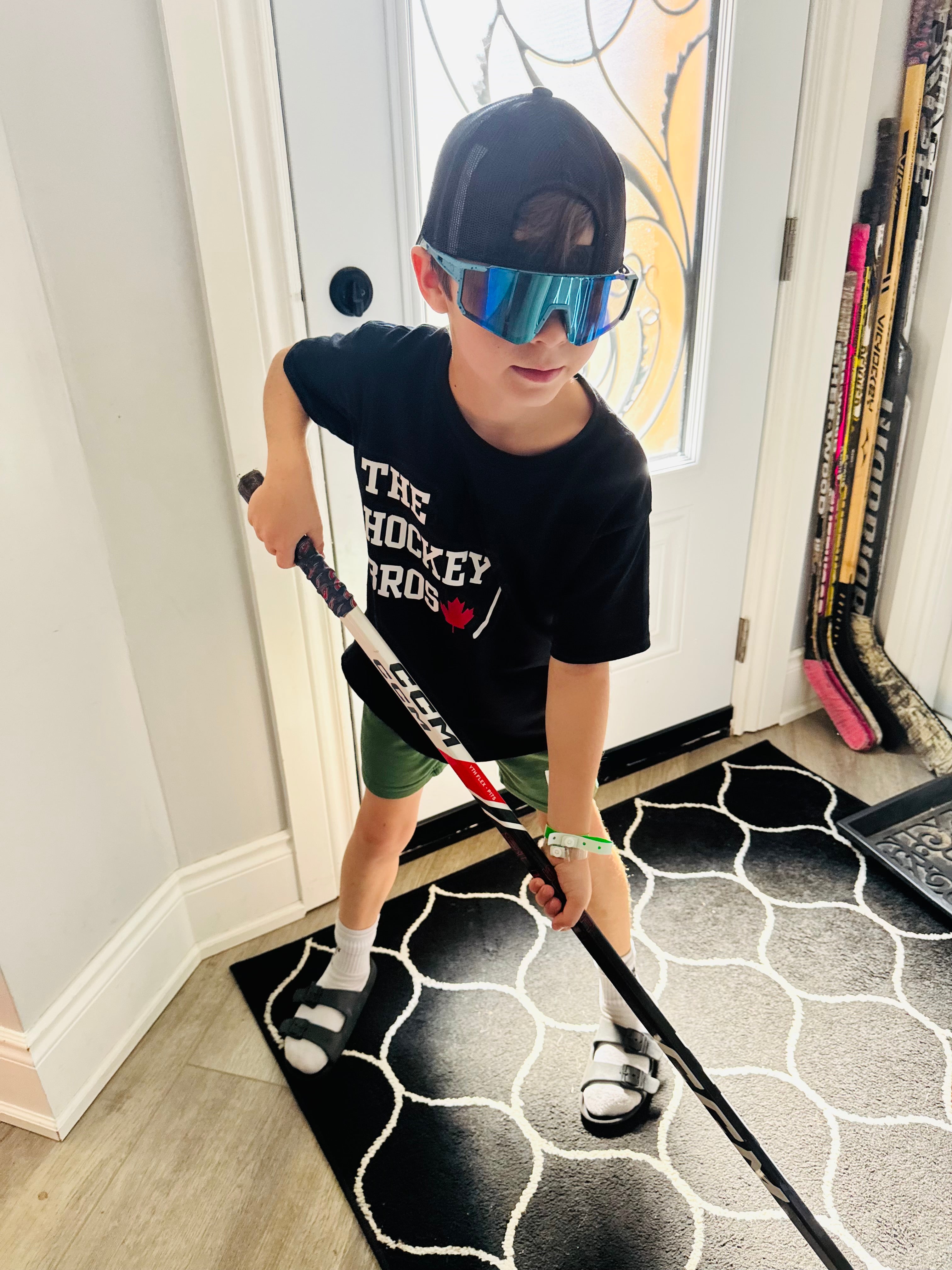 A child wearing a black graphic t-shirt, green shorts, a black cap, and reflective sunglasses holds a hockey stick