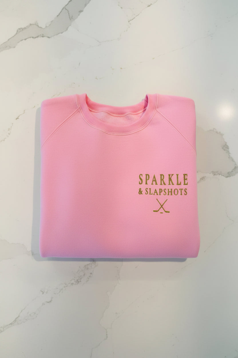 Women's pink crewneck sweatshirt with gold glitter vinyl Sparkle & Slapshots logo and crossed hockey sticks — left-chest placement