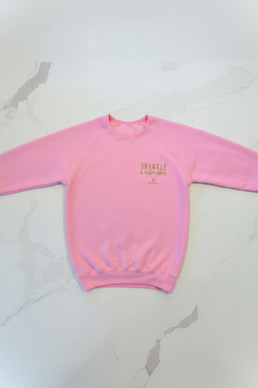 Close-up of gold glitter vinyl Sparkle & Slapshots crossed hockey sticks logo on pink crewneck sweater