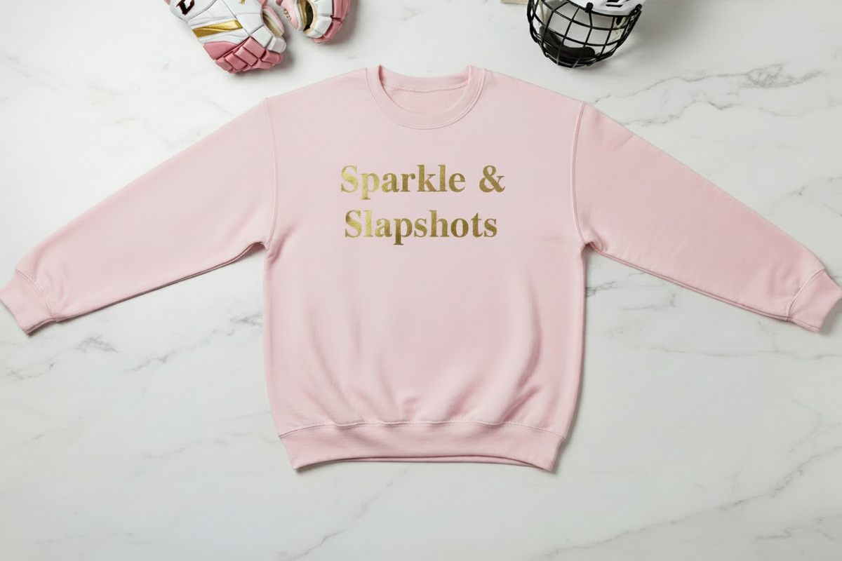 A pink crewneck sweatshirt with 'Sparkle & Slapshots' and crossed hockey sticks printed in gold on the front, laid flat on a white marble background. A black hockey puck is placed below the text.