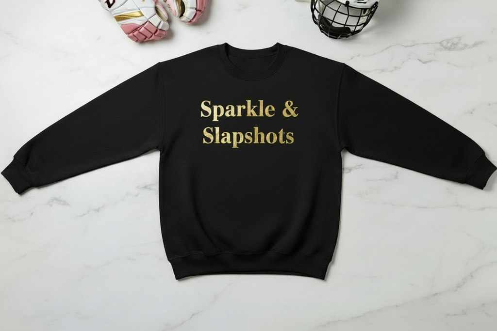 A black crewneck sweatshirt with 'Sparkle & Slapshots' printed in gold text across the chest, laid flat on a white marble background with hockey gear props.